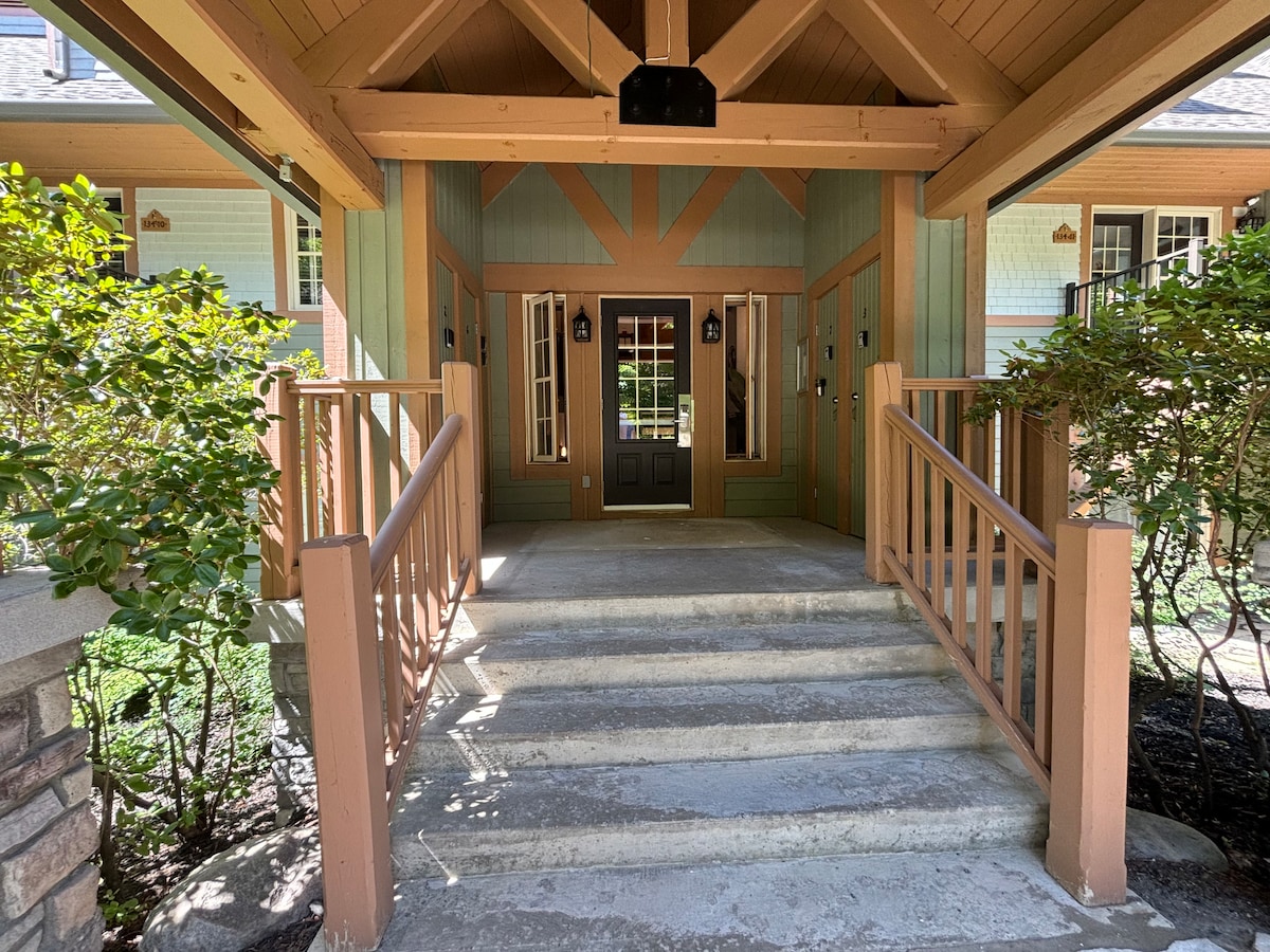 An inviting entrance features wide stone steps leading up to a wooden door framed by glass panels. Flanking the door are large windows, and railing lines the porch, surrounded by lush greenery that enhances the welcoming appeal of the entryway.