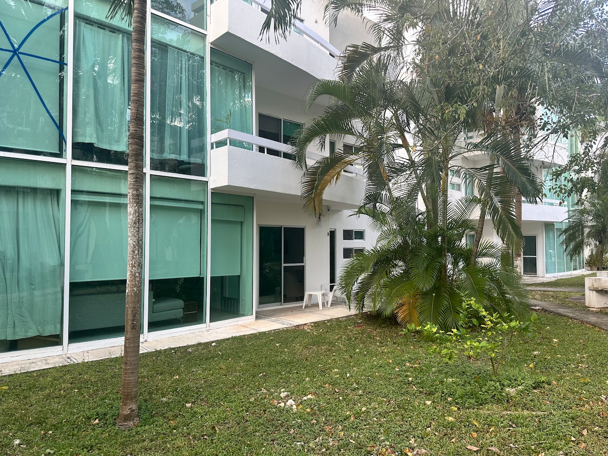 A modern building is surrounded by lush greenery and palm trees. Large glass windows provide abundant natural light to the open spaces. A small patio area features a few white chairs, offering a peaceful spot to relax amidst the natural setting.