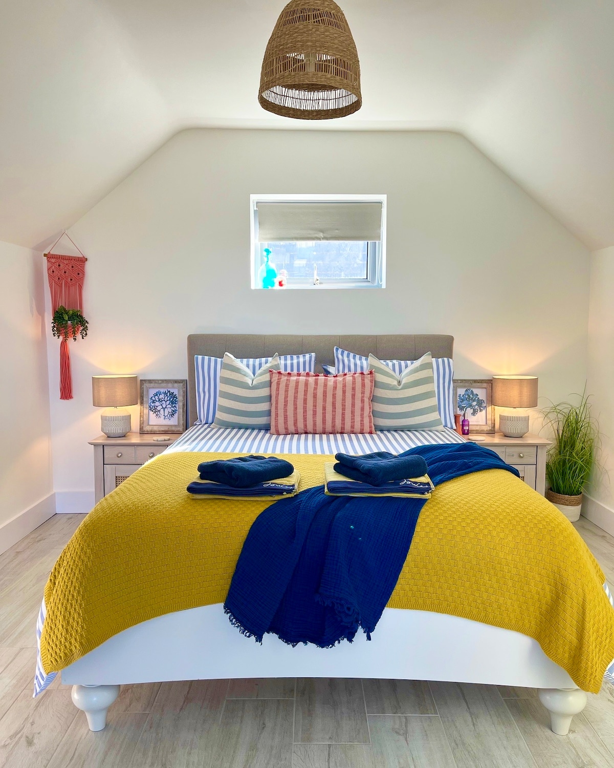 A sleeping area is highlighted by a spacious bed adorned with bright yellow bedding and coordinated throw blankets. Two bedside tables with lamps frame the bed, while decorative pillows in various patterns add visual interest. Natural light enters through a small window, enhancing the fresh atmosphere.