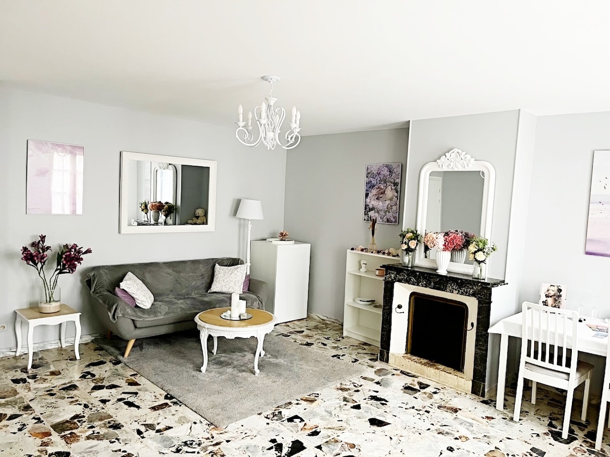 A well-arranged living area features a gray sofa and a round coffee table, complemented by a plush rug. Light-colored walls and decorative art enhance the space, while a chandelier and mirror add elegance. A mantelpiece holds floral decor, providing a cozy yet refined atmosphere.