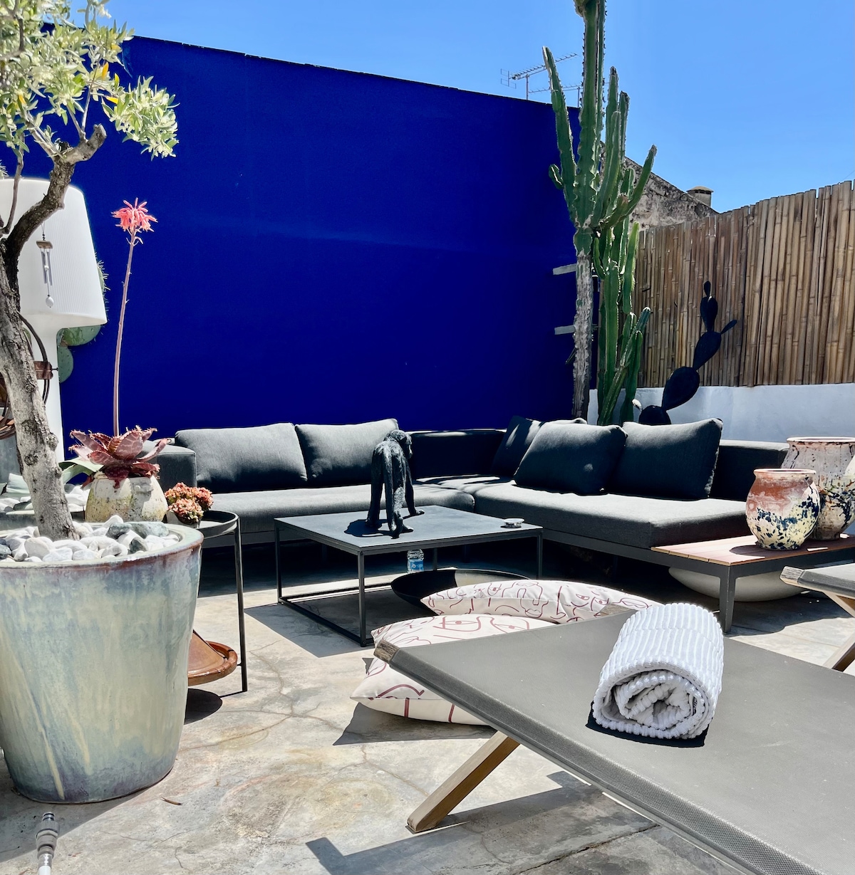 An outdoor seating area is composed of a black sectional sofa and low tables. Potted plants and decorative sculptures add interest, while a vibrant blue wall contrasts with the earthy tones of the stone floor. Cushions are neatly arranged for comfort.