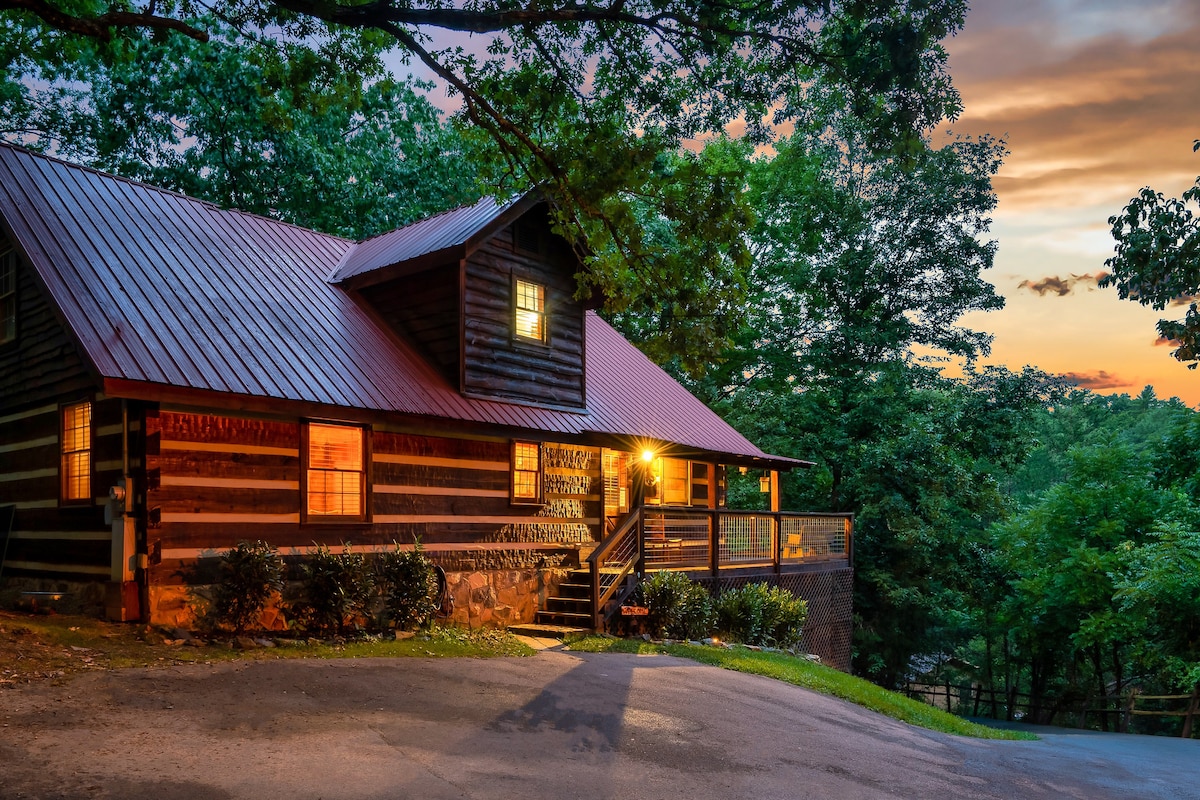A charming cabin is set among lush trees, featuring a rustic wooden exterior with a red metal roof. Warm lights glow from the windows, inviting guests to explore. The wraparound deck is visible, providing a cozy outdoor space for relaxation. The sunset casts a soft hue in the background.