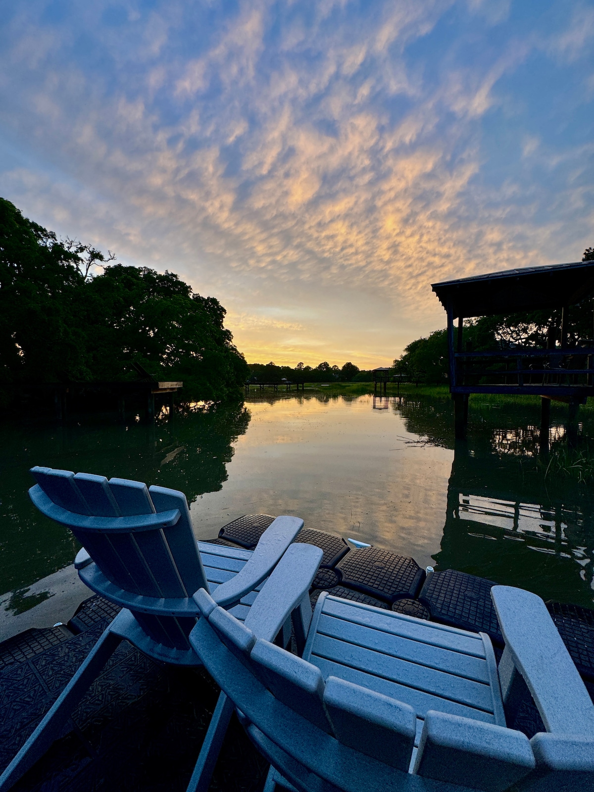 Two light blue outdoor chairs are positioned on a dock overlooking a calm tidal creek. The water reflects the pastel hues of the sunset sky, while surrounding trees frame the peaceful scene. The atmosphere suggests a tranquil setting for relaxation and nature observation.