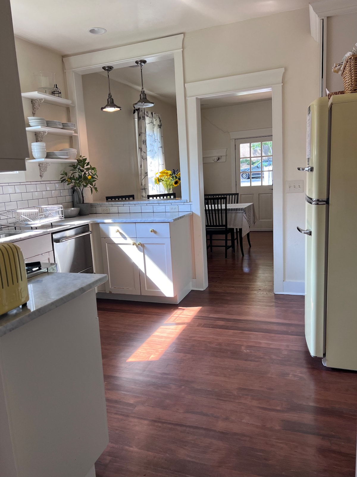 An inviting kitchen features modern appliances, including a retro yellow refrigerator and a sleek silver dishwasher. Natural light fills the space, highlighting the white cabinetry and Carrara marble countertops. A dining area is visible through the doorway, furnished with a table and black chairs.