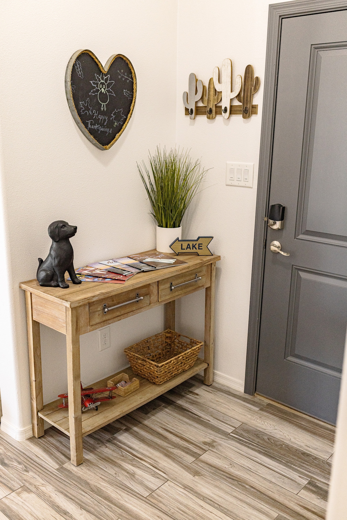 An entryway table made of light wood is positioned against a wall. It features two drawers, a container for magazines, and a decorative black dog figurine. Above, a chalkboard in a heart shape and wall decor in the form of cacti are displayed.