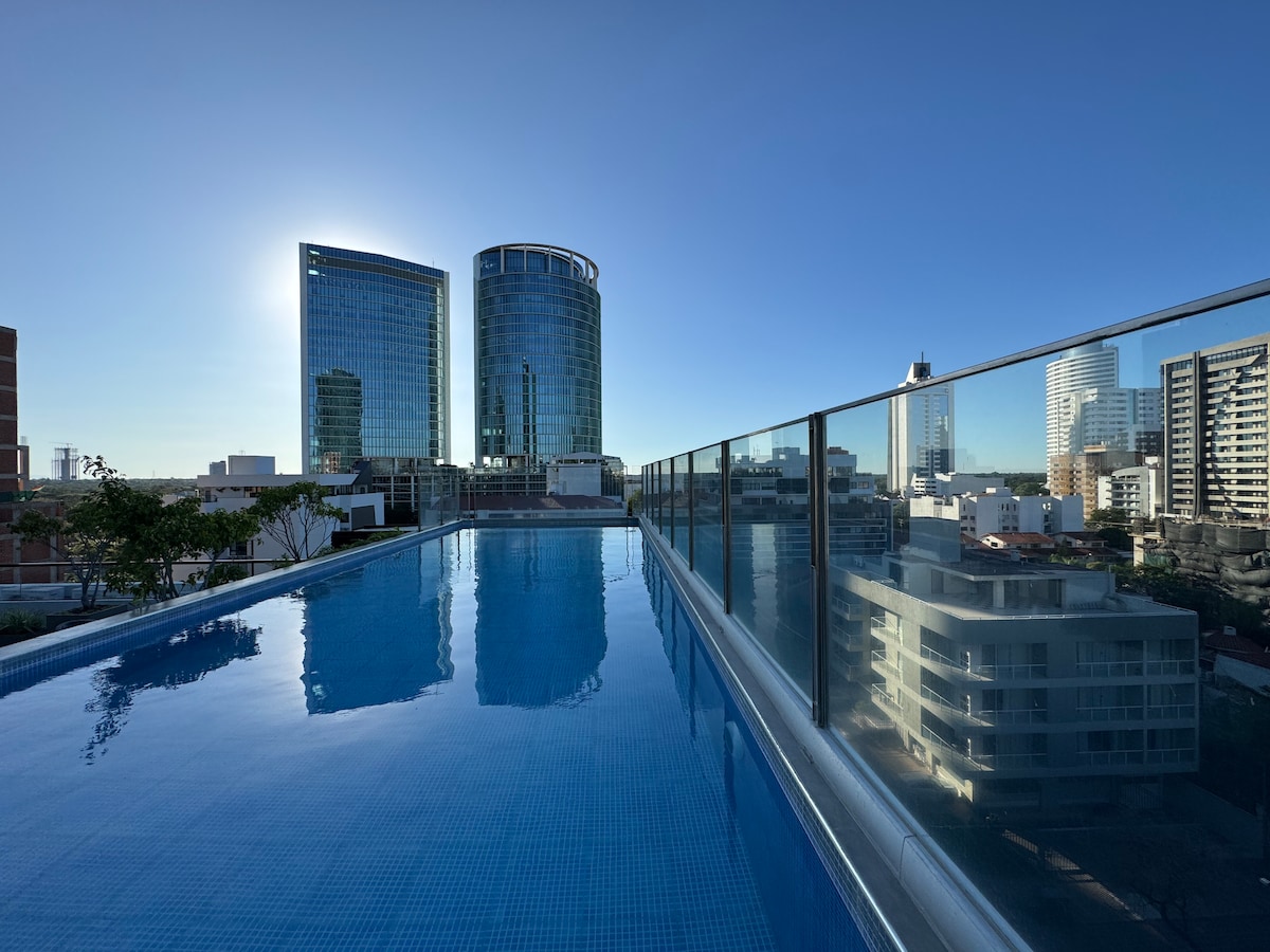 A rooftop swimming pool reflects the clear blue sky, offering views of contemporary high-rise buildings. The pool area is surrounded by a glass railing, emphasizing an open and spacious atmosphere. Urban scenery is visible in the background, characterized by a mix of modern architecture.