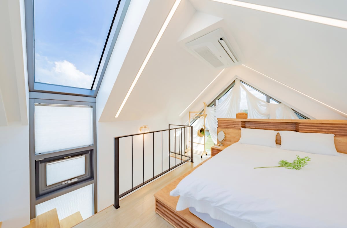 A bright loft space is features a spacious bed with wooden accents, complemented by large windows that invite natural light. Sleek architectural lines enhance the airy feel, while a small green plant adds a touch of nature to the serene atmosphere.