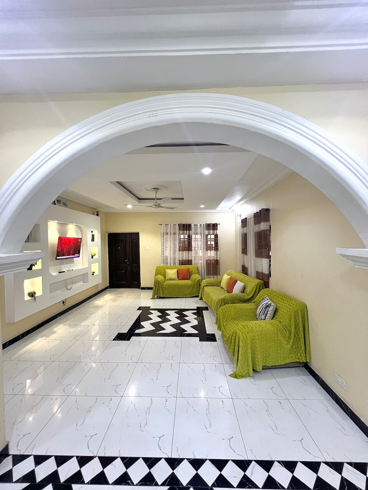 A welcoming living room features a curved archway leading into a bright space. Two green sofas adorned with accent pillows face a wall-mounted television. Natural light enters through the sheer curtains beside a central geometric rug on polished tile flooring.