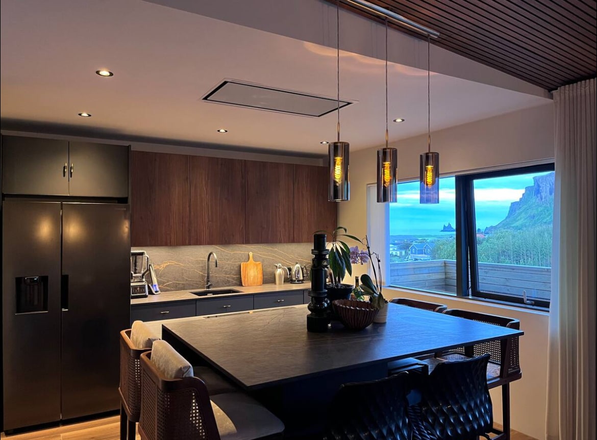 A modern kitchen features a large kitchen island equipped for four diners. Pendant lights hang above, illuminating the dark cabinetry and ample counter space. A window provides a view of the outdoors, enhancing the space's brightness with natural light.