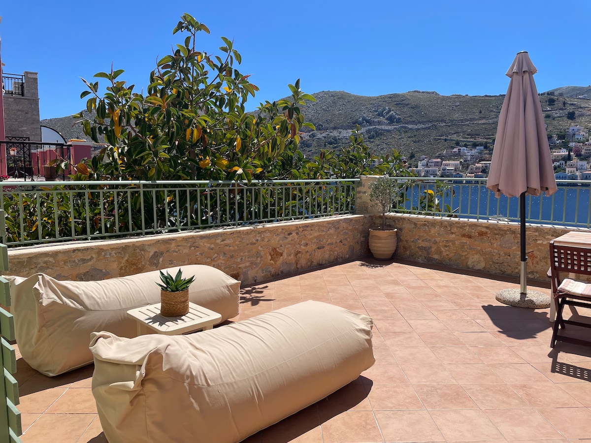 An outdoor terrace features two large bean bag chairs and a small wooden table. A potted plant sits on the table, surrounded by a stone wall. A large umbrella provides shade, with scenic views of the coastline visible in the background.