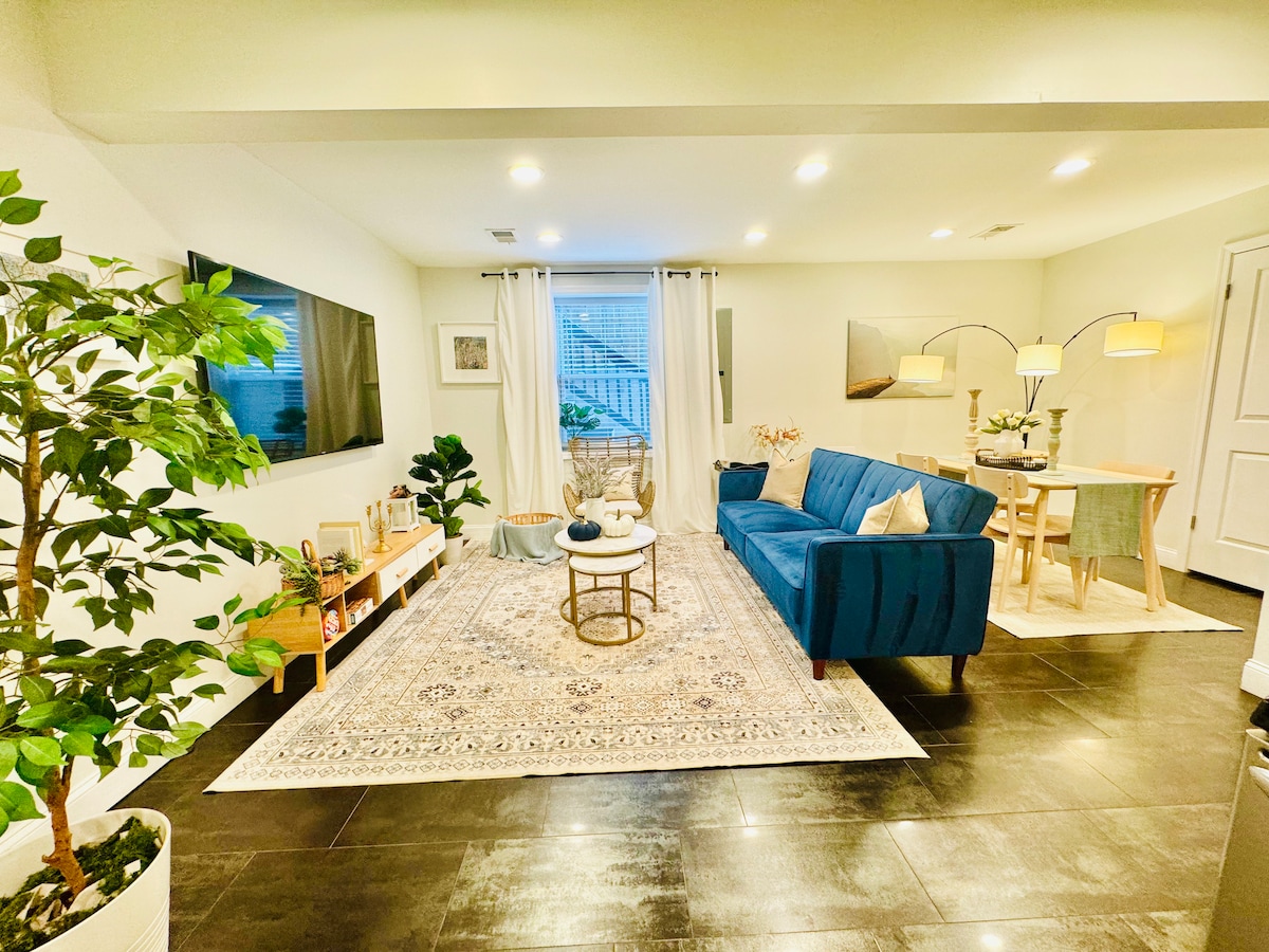A cozy living area is highlighted by a blue sofa and a round coffee table placed on an area rug. A flat-screen TV is mounted on the wall opposite the sofa. A small dining table with two chairs is visible near the window, complemented by floor lamps and indoor plants.