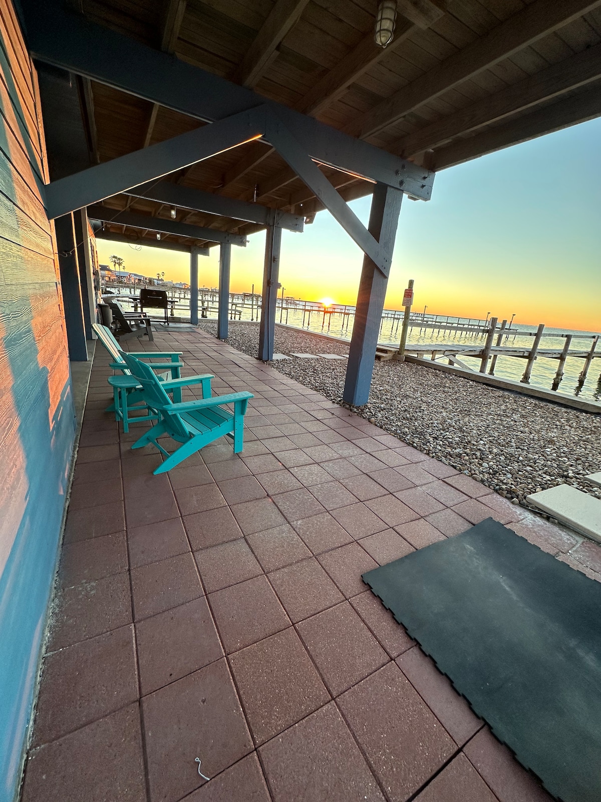 A serene patio offers two turquoise adirondack chairs, situated along a tiled walkway. The setting sun casts warm hues over the scene, while a private fishing pier extends into the water, creating a backdrop of tranquility.
