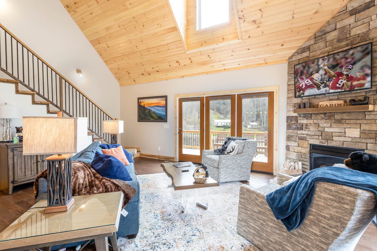 A welcoming great room features vaulted wood ceilings and large windows that bring in natural light. Comfortable seating arrangements surround a coffee table, with a stone fireplace and a mounted television enhancing the space. A blend of modern and rustic decor complements the atmosphere.