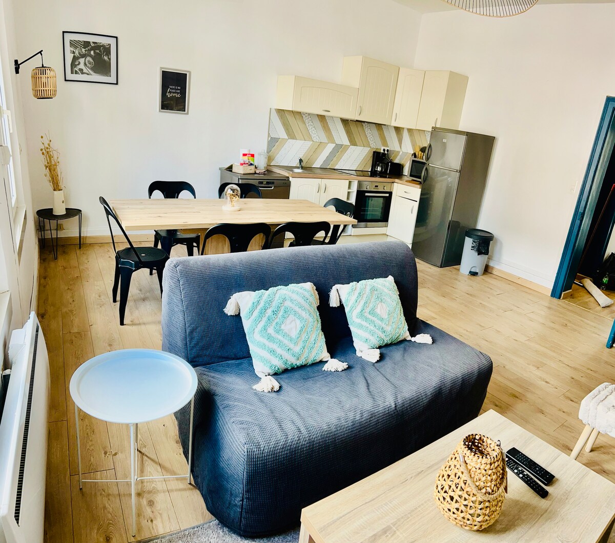 An open living area features a comfortable sofa with decorative cushions, a dining table with black chairs, and a well-equipped kitchen area in the background. Light-colored cabinetry and appliances create a spacious feel, while natural wood flooring enhances the warm ambiance.
