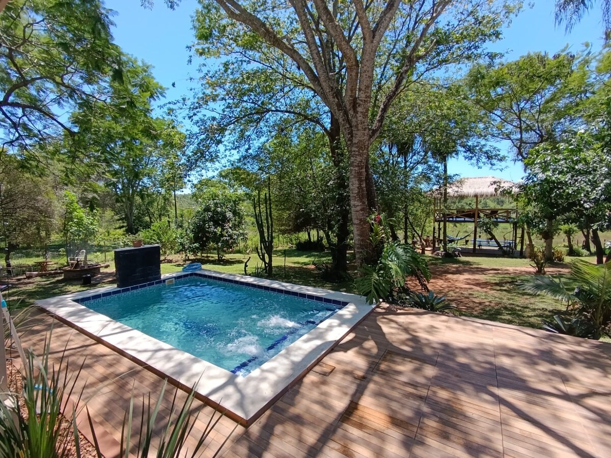 A private outdoor area features a sparkling pool surrounded by wooden decking. Lush greenery and tall trees create a serene atmosphere, while a shaded seating area complements the natural setting. In the background, a children's play area is visible, enhancing the family-friendly environment.