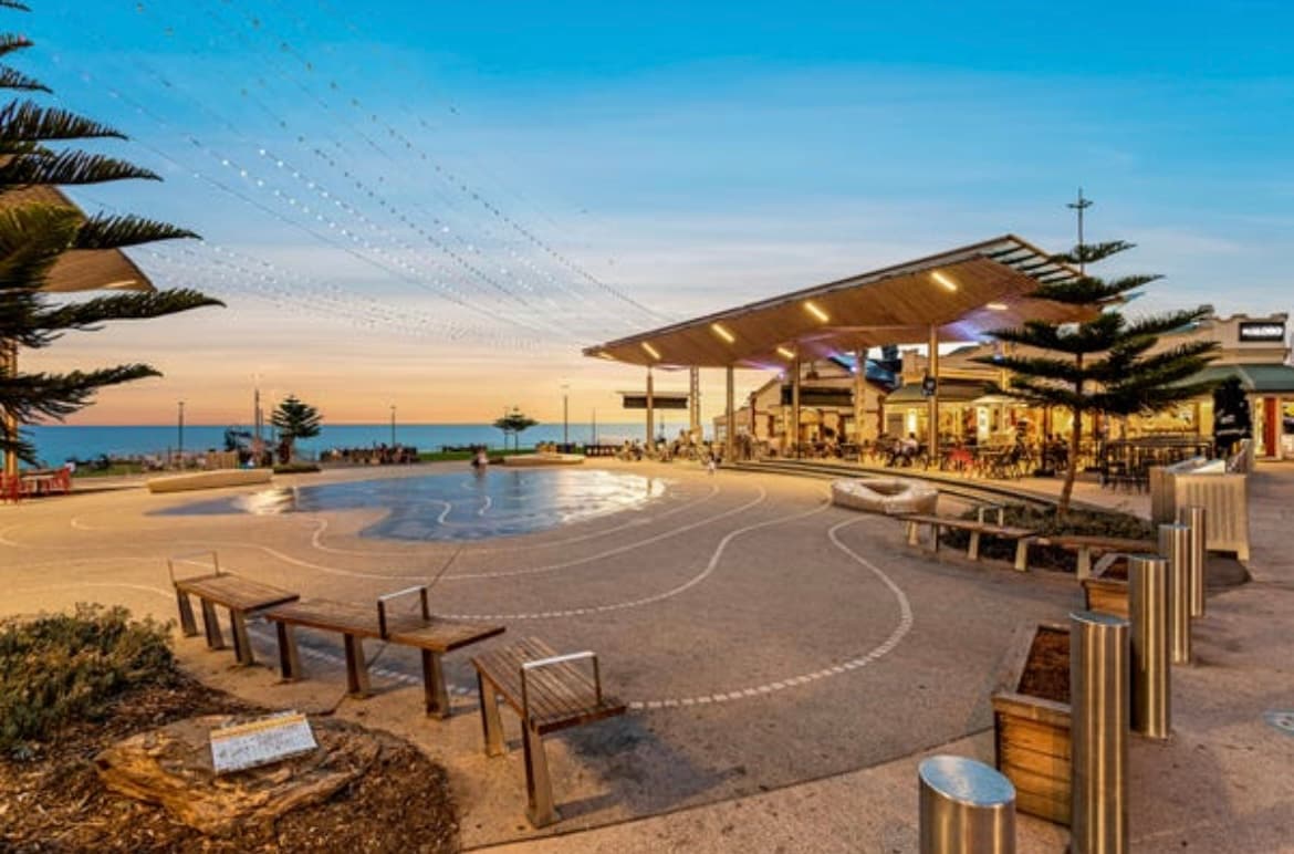 An outdoor area is depicted with a smooth, expansive concrete surface and multiple seating options. Subtle paths are drawn in white. A structure with a roof offers shade, surrounded by palm trees. The ocean sparkles in the background under a colorful sunset sky.