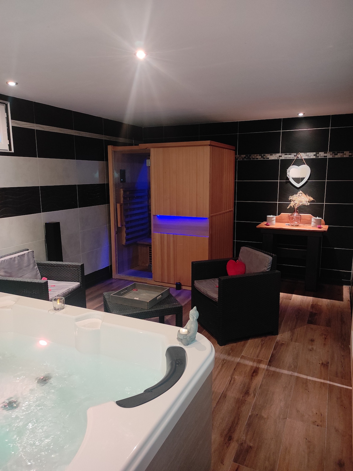 A calming wellness area features a jacuzzi alongside a sauna, with soft lighting highlighting the space. Two wicker chairs invite relaxation, while decorative elements add a touch of warmth. The wood paneling of the sauna contrasts with the modern tiled walls.