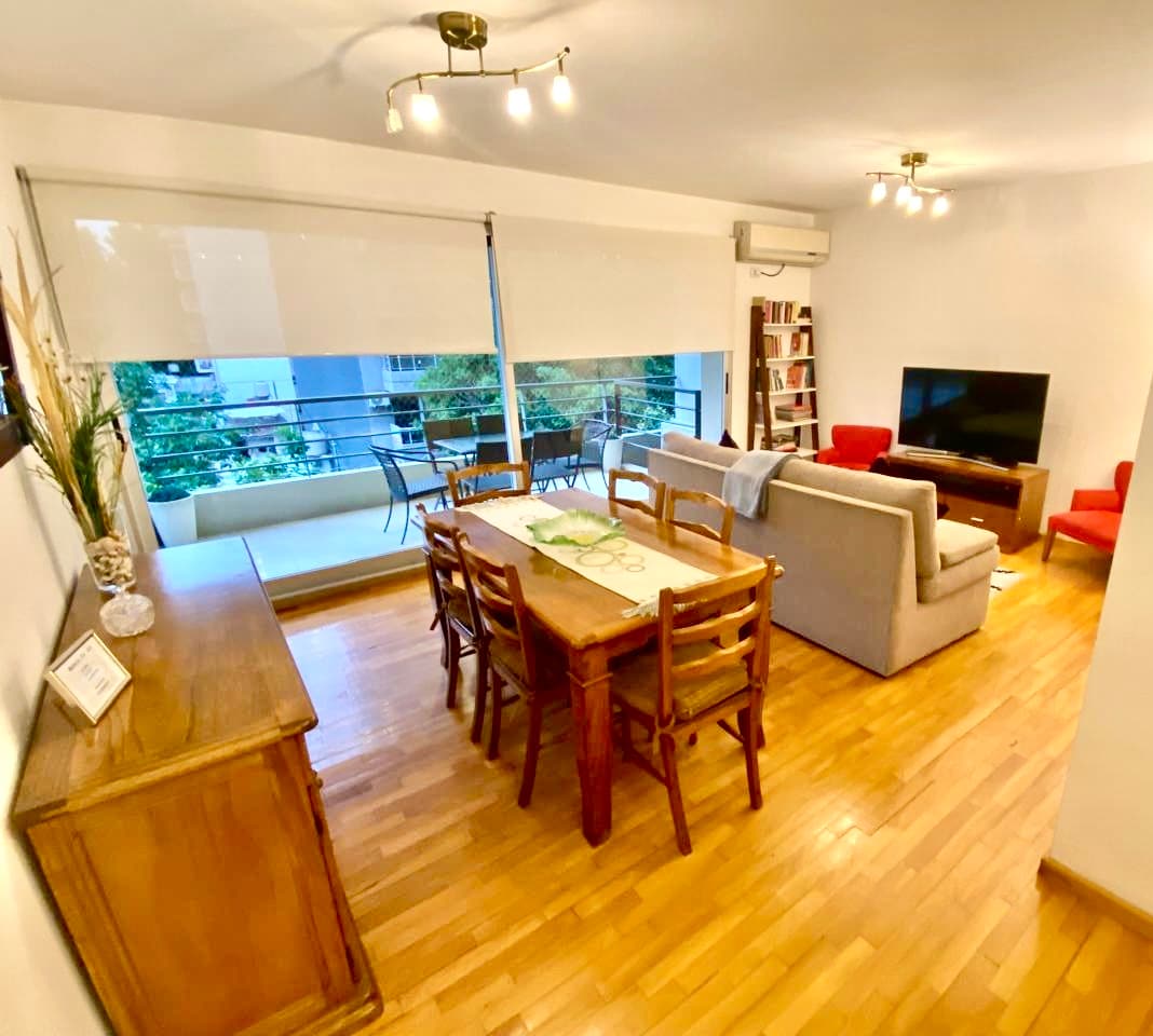 The open living and dining area features a wooden dining table with six chairs, a comfortable couch, and two accent chairs. A large window provides natural light and views of a balcony with outdoor seating. A Smart TV is positioned on a media unit, while decorative elements are visible.