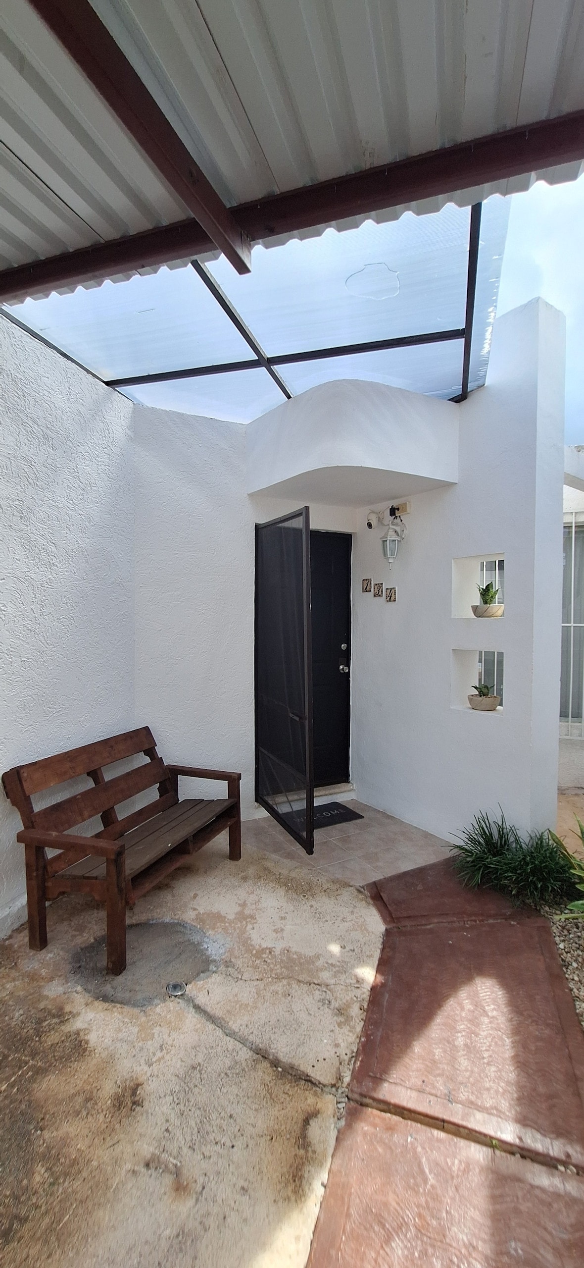The entrance features a modern design with a wooden bench beside the door. A glass roof allows natural light to fill the space. Clean white walls provide a bright, open feel, while small built-in niches display decorative plants.