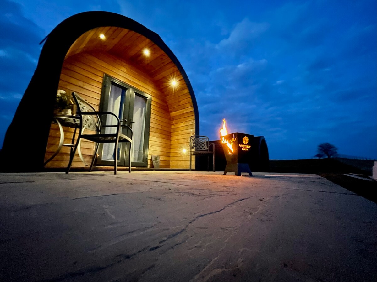 A cozy cabin exterior is illuminated by soft lighting at dusk. A seating area with two chairs is positioned next to a black fire pit, which displays a small flame. The smooth surface of the patio contrasts with the wooden cabin structure.