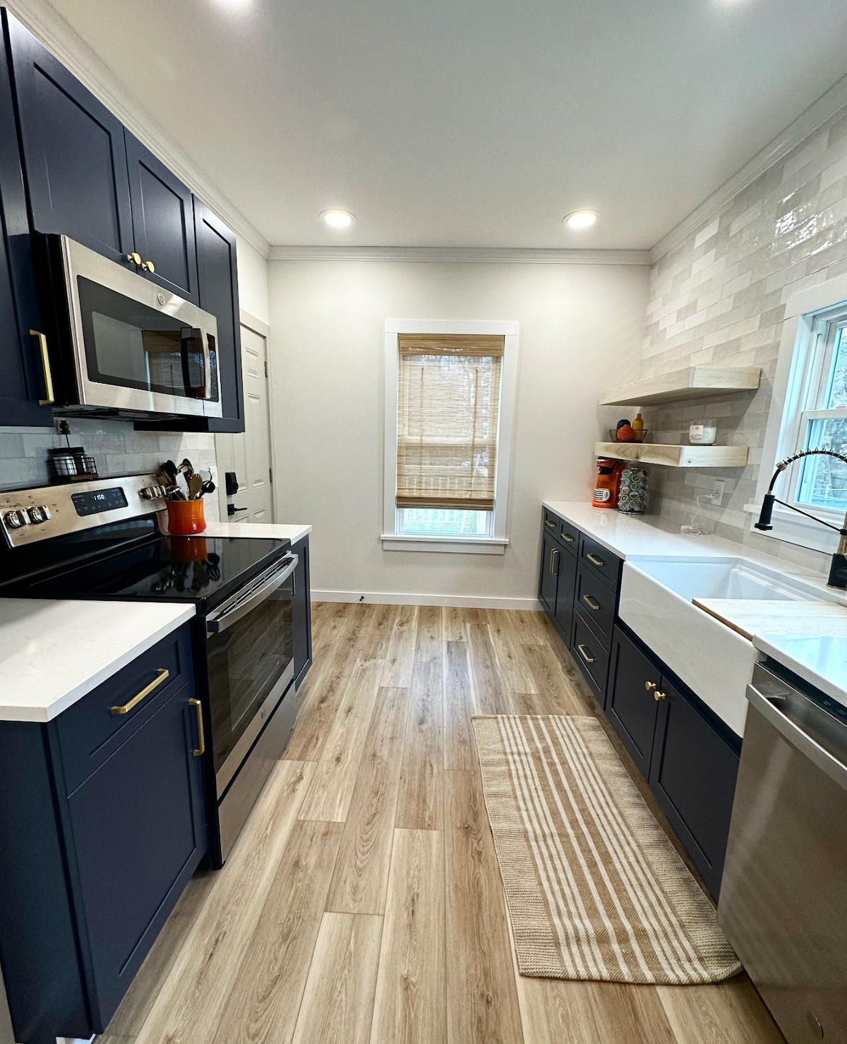 The kitchen features modern cabinetry in a deep navy color, complemented by white countertops and tile backsplash. Stainless steel appliances include an oven, microwave, and dishwasher. A window allows natural light into the space, and a patterned rug lies on the floor.