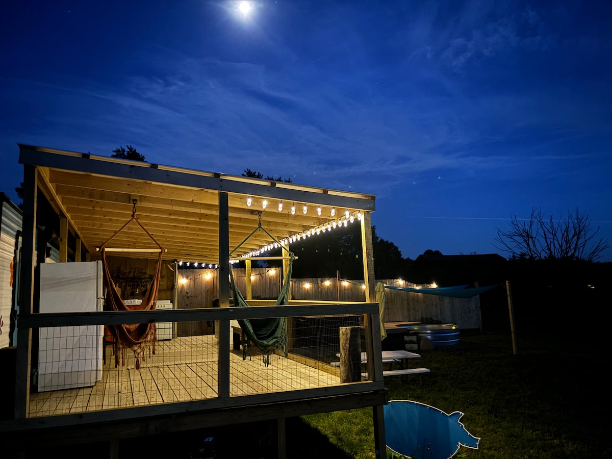 A wooden deck illuminated by string lights creates an inviting atmosphere at night. Two hammocks hang from the rafters, and an area for outdoor seating is visible. A stock tank pool is situated in the yard, reflecting the moonlight against a backdrop of lush greenery.