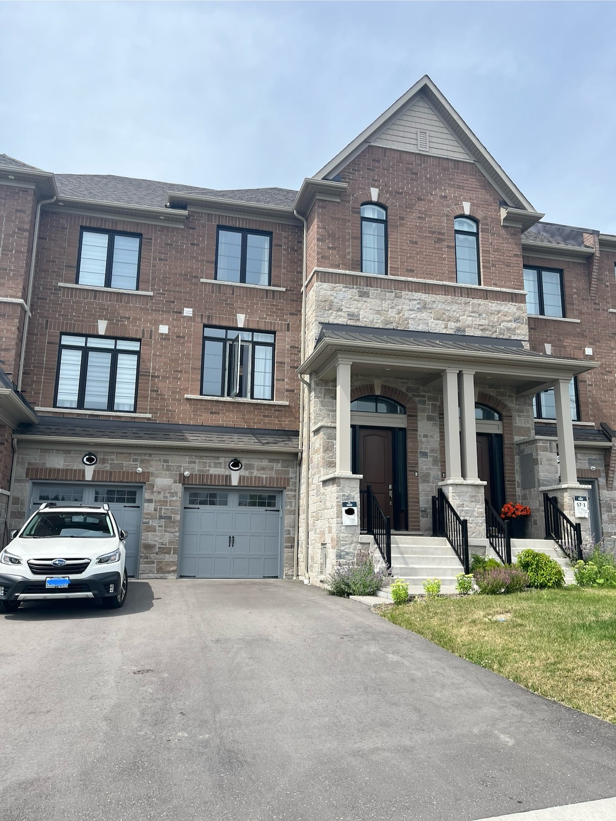 The exterior of a multi-level brick home is captured, highlighted by large windows and a prominent covered entryway. A well-maintained driveway leads to an attached garage, with a vehicle positioned in front. Lush greenery is visible along the sides of the property.