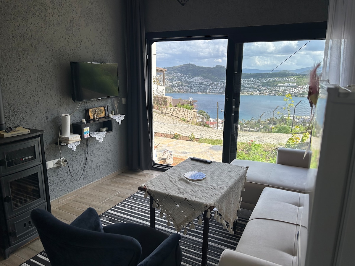 A comfortable living space features a spacious white sofa and a cozy black armchair, set against a backdrop of expansive glass doors revealing a stunning sea view. A simple table is positioned in front, with a decorative plate resting on it. Natural light streams into the room.