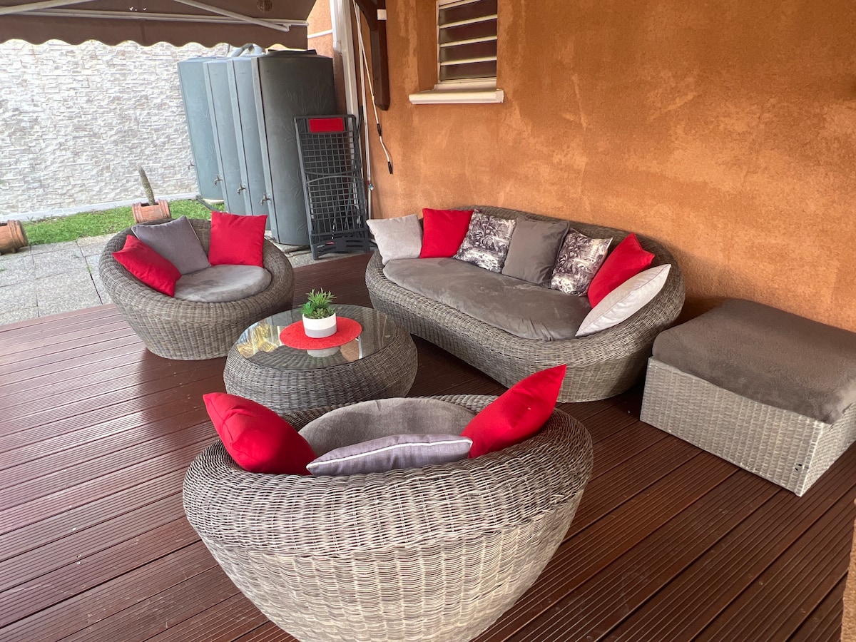 A cozy outdoor seating area features a woven rattan sofa, adorned with red and gray pillows. A round coffee table rests in the center, accompanied by a small potted plant. The space is surrounded by a wooden deck, providing a comfortable spot for relaxation.
