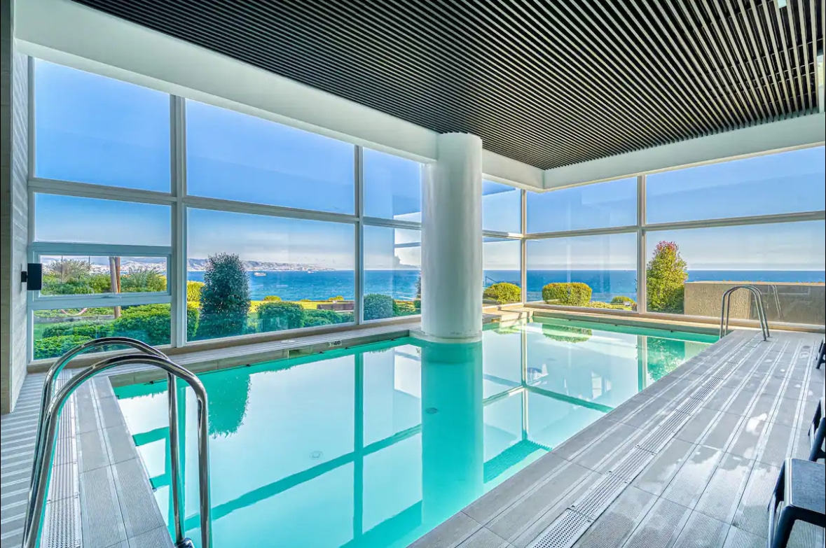 A bright indoor pool area is highlighted by large glass windows offering panoramic views of the ocean. The pool is surrounded by a sleek wooden deck, and well-maintained greenery is visible outside, creating a serene setting.