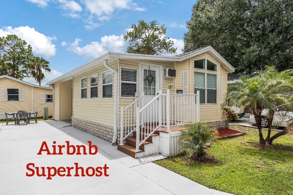 A charming cottage exterior is displayed with light yellow siding and large windows allowing natural light. A welcoming porch leads up to the entrance, while landscaped greenery and a palm tree enhance the surrounding area. A driveway provides convenient parking.
