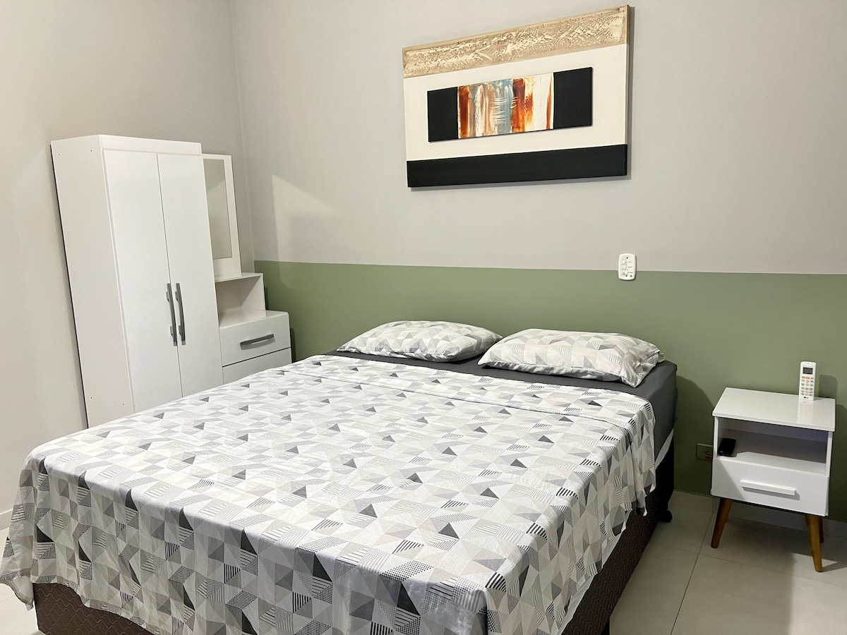 A spacious sleeping area is furnished with a queen bed featuring light-coloured bedding and two pillows. A simple nightstand rests beside the bed, while a white cabinet is positioned against the wall. Art hangs above the bed, adding visual interest to the room.