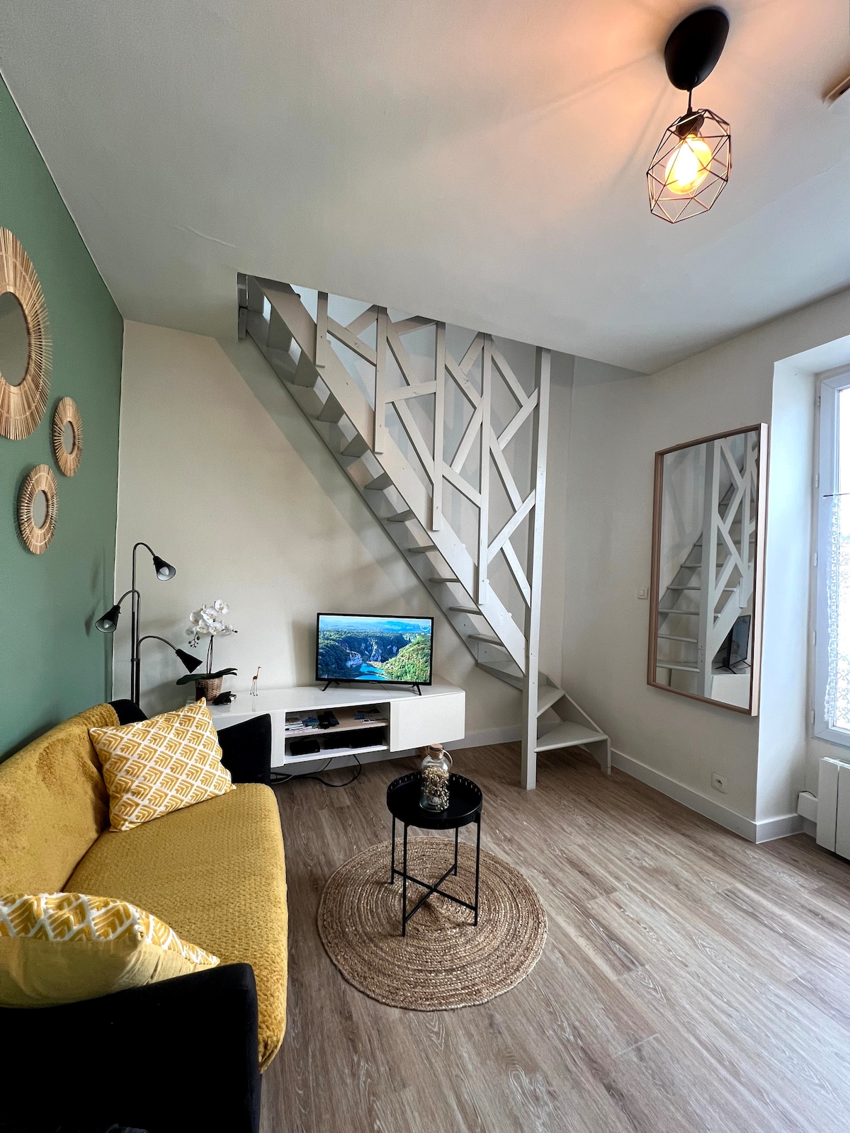 A living area showcases a yellow sofa adorned with a patterned cushion. A small black table is centered on a round rug. A television is positioned on a low white cabinet, while a staircase leading to the upper level is visible in the background. Natural light filters through a nearby window.