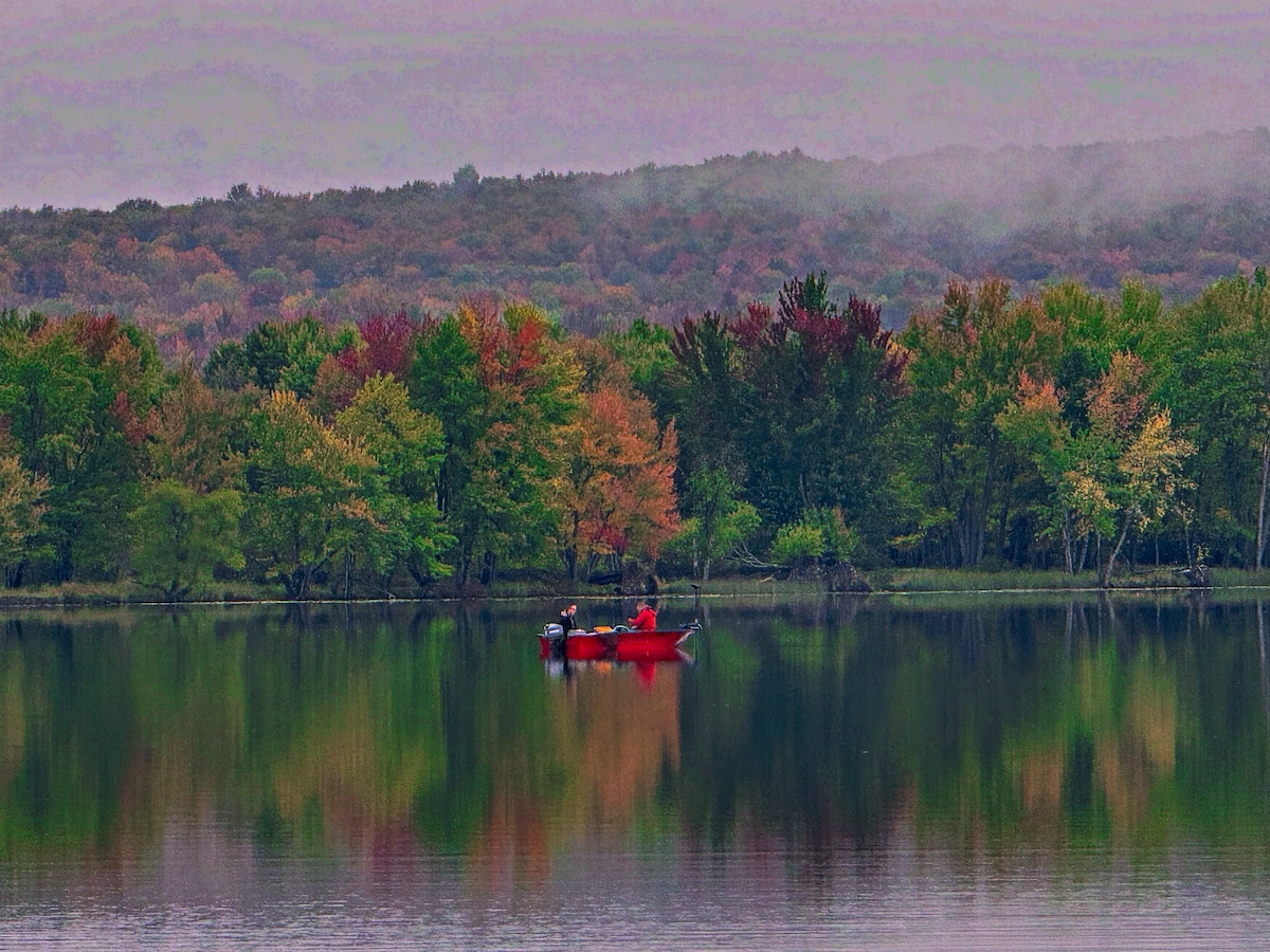 A still lake reflects the autumnal hues of the surrounding trees, ranging from greens to vibrant reds and oranges. A small red boat is seen floating gently on the water, contributing to the serene atmosphere of the scene.