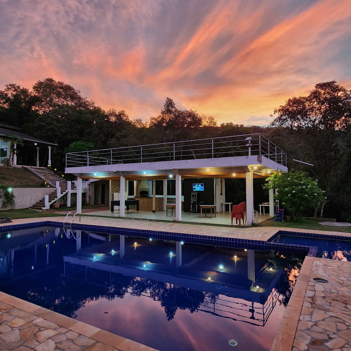 The spacious outdoor area features a large swimming pool reflecting a colorful sunset. Surrounded by greenery, the modern house offers an elevated terrace, with multiple seating options visible. The warm evening light enhances the tranquil setting, perfect for relaxation.