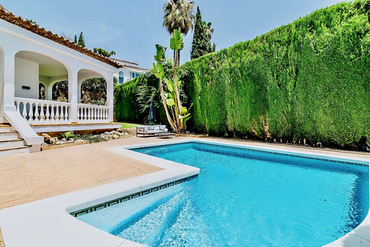 A private heated pool is surrounded by manicured hedges, providing an inviting space for relaxation. The pool deck features sun loungers and a tiled border, while a covered terrace is visible in the background, enhancing the outdoor living experience.