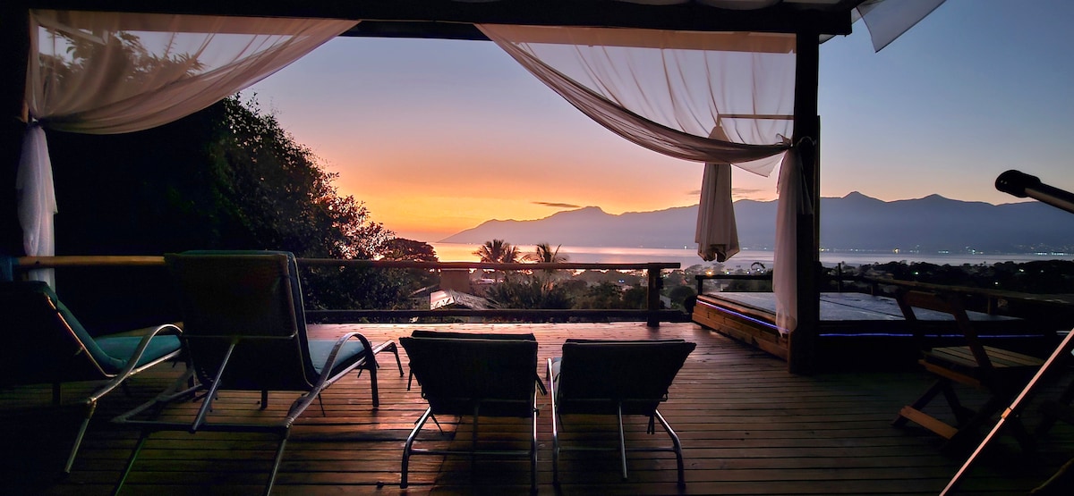 A spacious deck features two lounge chairs, facing a serene view of the sunset over distant mountains and the ocean. Sheer curtains billow softly in the evening breeze, enhancing the peaceful atmosphere of the outdoor space, which includes a jacuzzi in the background.