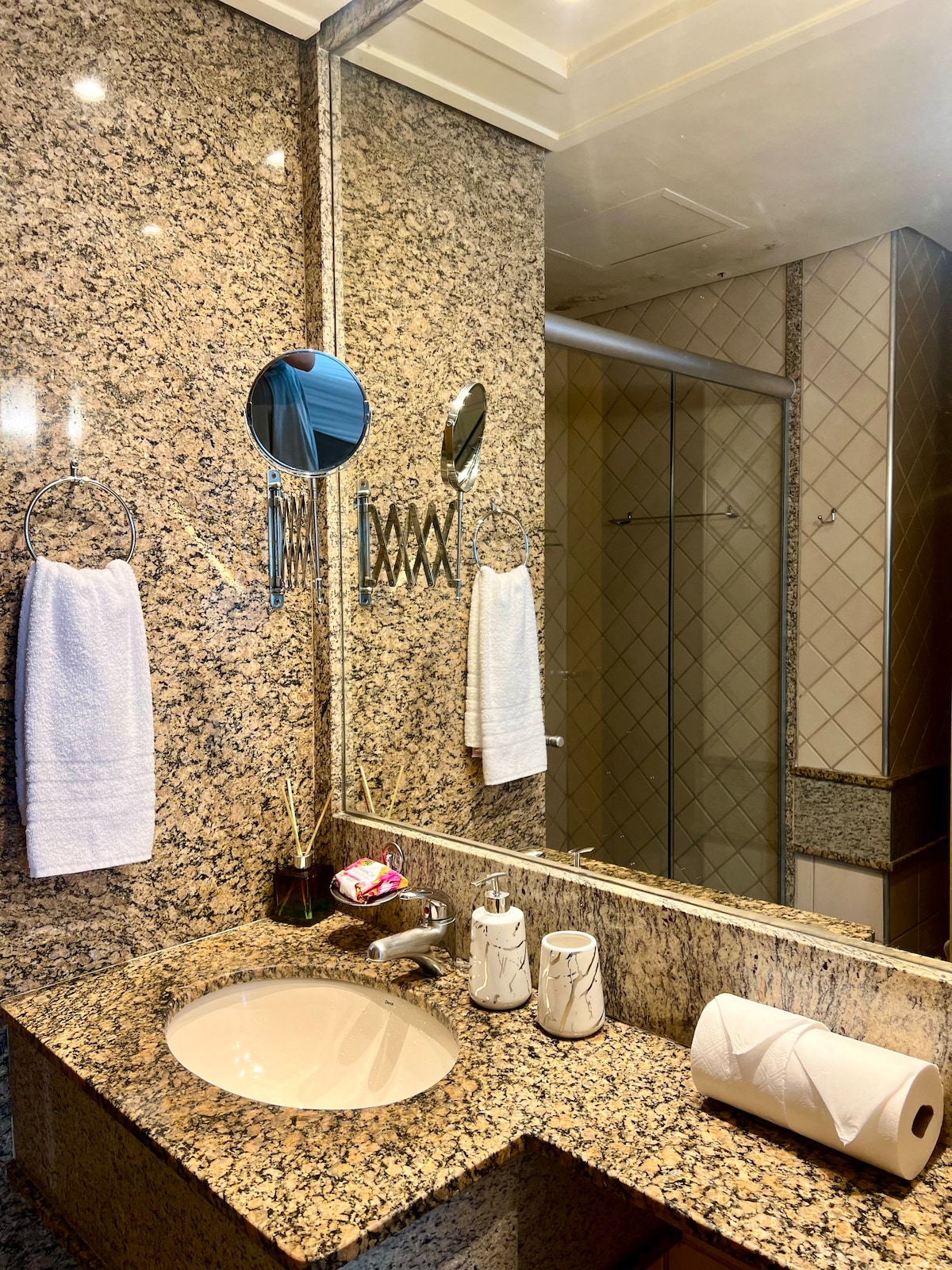 A bathroom features a granite countertop with a sink and mirror. Hand towels are neatly hung on towel rings. Decorative items include a soap dispenser and a tissue roll displayed on the counter. The tiled walls and shower area are visible in the background.