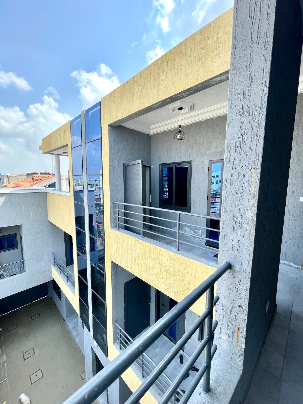The exterior of a modern building is observed, featuring a combination of yellow and gray walls. Large windows allow natural light to enter, and a balcony with a railing is visible, enhancing the architectural design. The surroundings are bright with clear skies above.