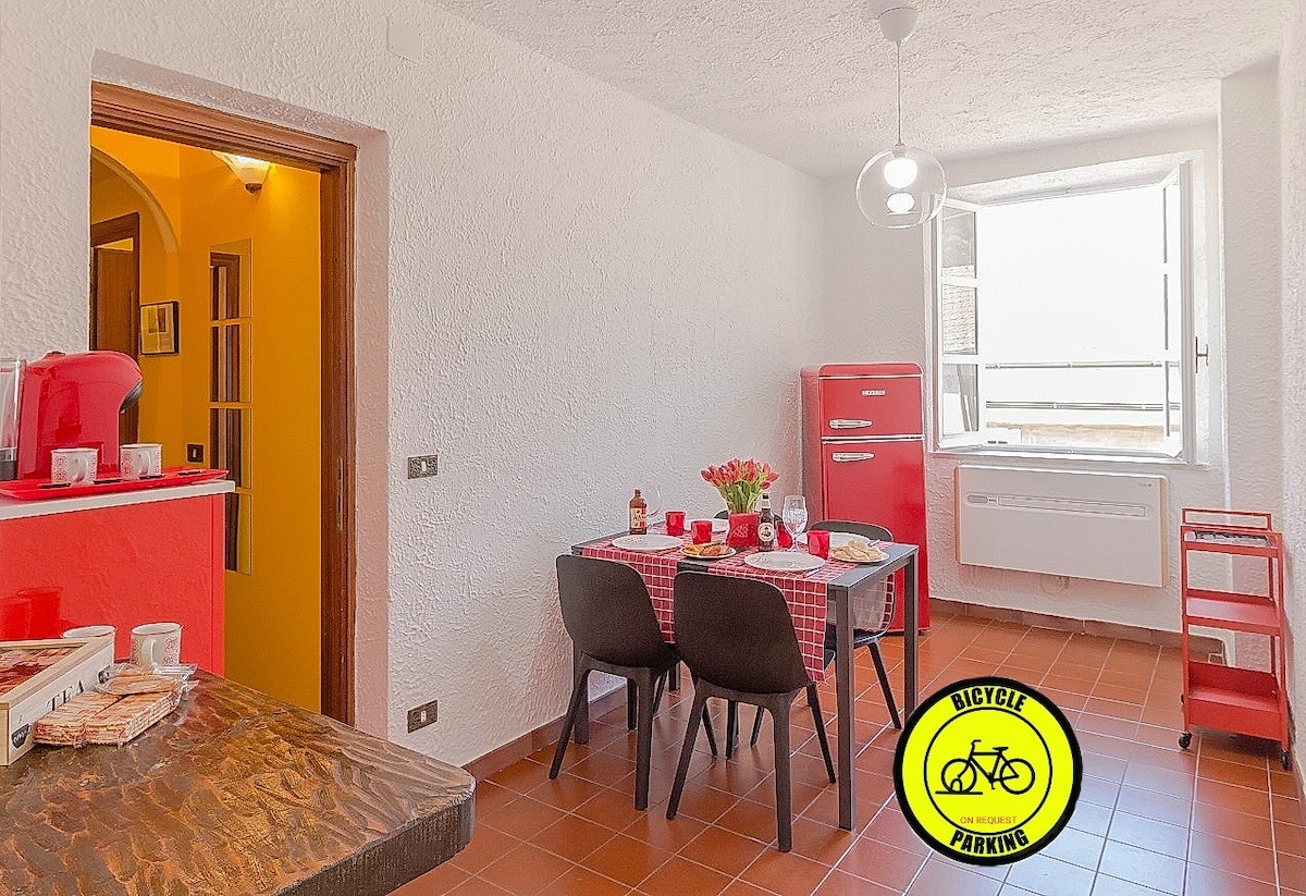 A modern kitchen and dining area are visible, featuring a red refrigerator and a dining table set for four. Natural light enters through the window, illuminating the room's white walls and terracotta floor. A bicycle parking sign is prominently displayed.