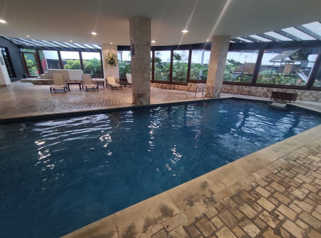 An indoor pool area is highlighted by large windows allowing natural light. The inviting blue water is surrounded by a stone deck, with a few lounge chairs placed nearby for relaxation amidst a lush garden view.