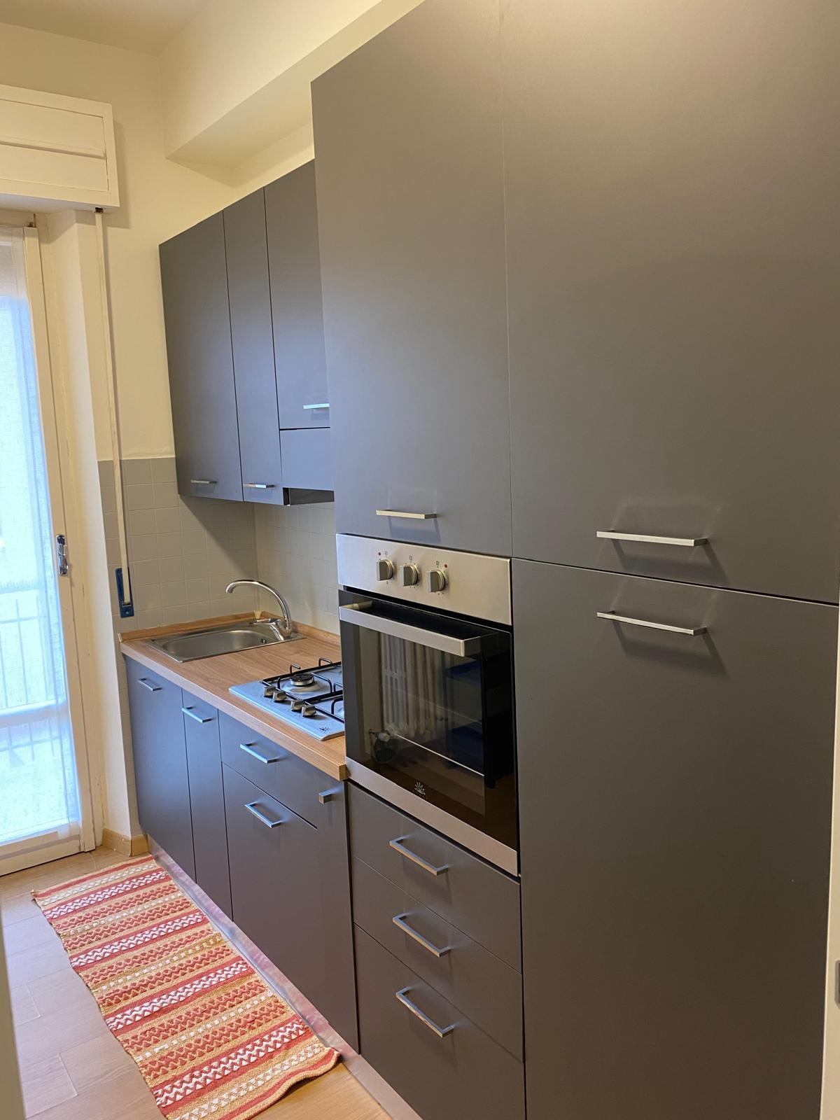 A modern kitchen is equipped with sleek gray cabinets and a wooden countertop. Cooking appliances include a gas stove and an oven, with a microwave mounted above. A small rug adds a touch of comfort in front of the sink area, while natural light enters through the adjacent window.