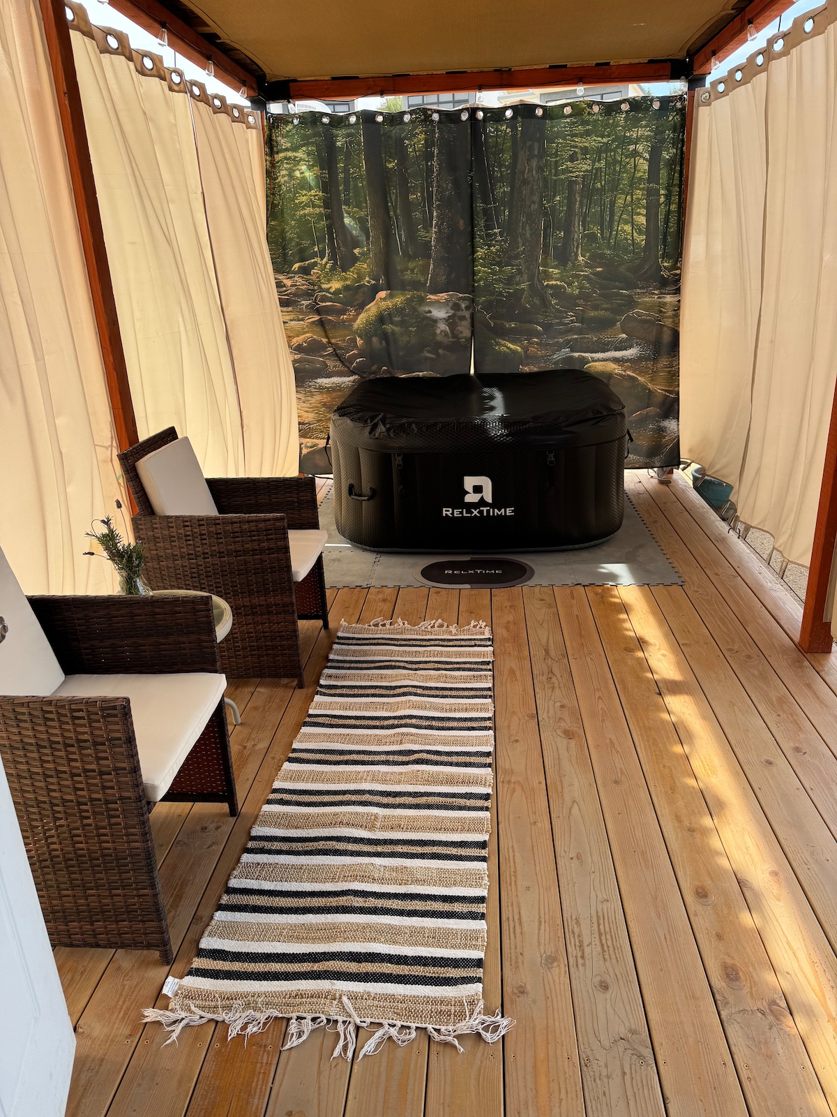A private space features a black jacuzzi surrounded by wood-paneled walls and a soft, natural-coloured floor. Two woven chairs are positioned on a striped rug, offering a cozy seating area. Soft curtains frame the area, adding an element of privacy and comfort.
