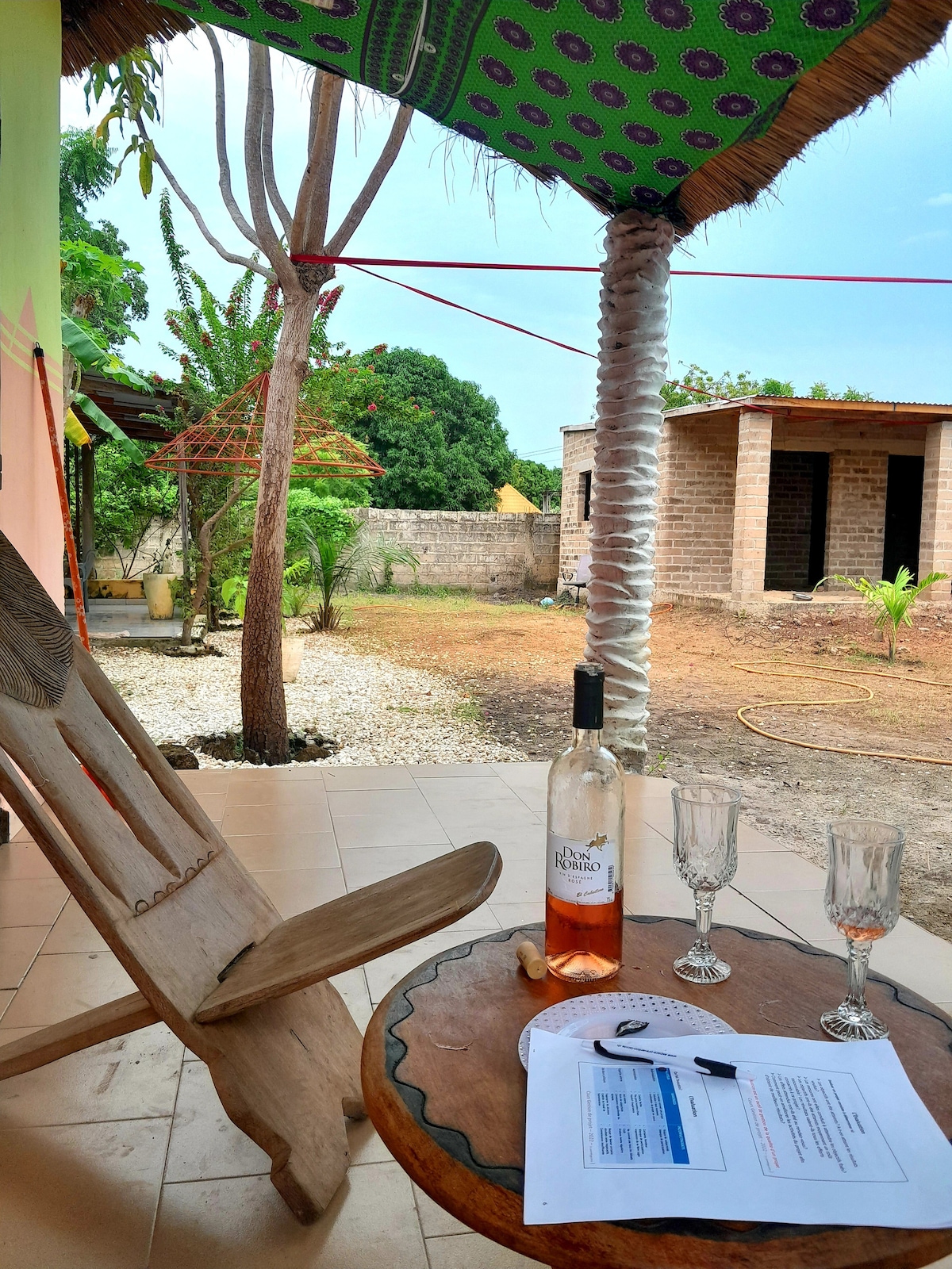 An outdoor seating area is presented, featuring a wooden chair placed beside a small round table. A bottle of wine and two glasses are arranged on the table. The view includes a garden space with greenery and a pathway leading to a nearby structure.