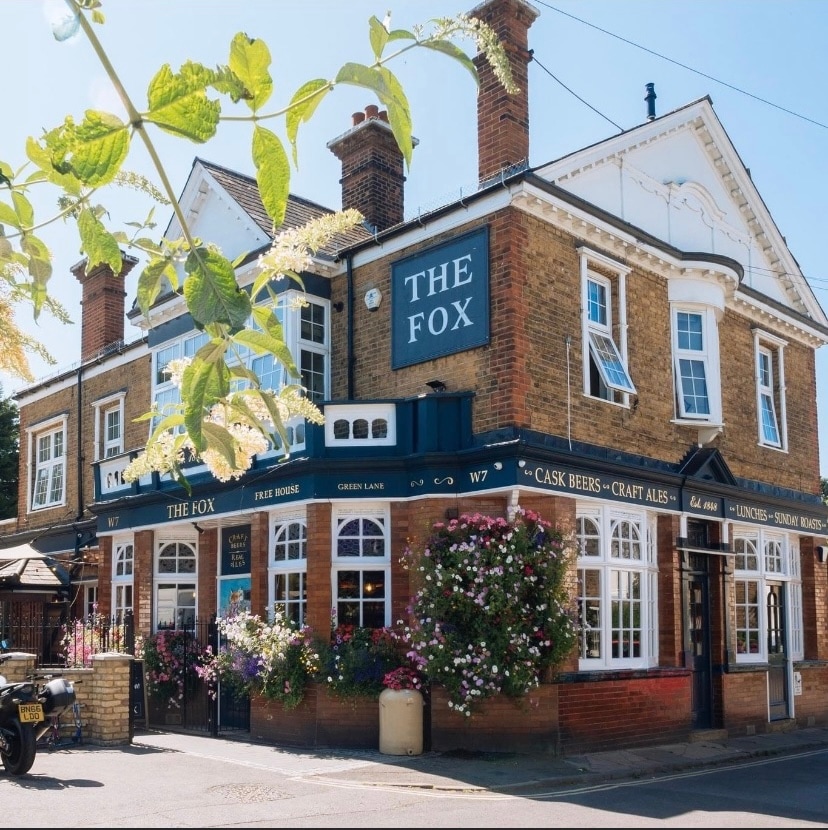 The exterior of The Fox Pub is displayed, featuring a traditional brick facade with large windows and vibrant flower displays. The signage is clearly visible, showcasing the establishment's name, while outdoor seating can be seen along the entrance, inviting guests to enjoy the surroundings.