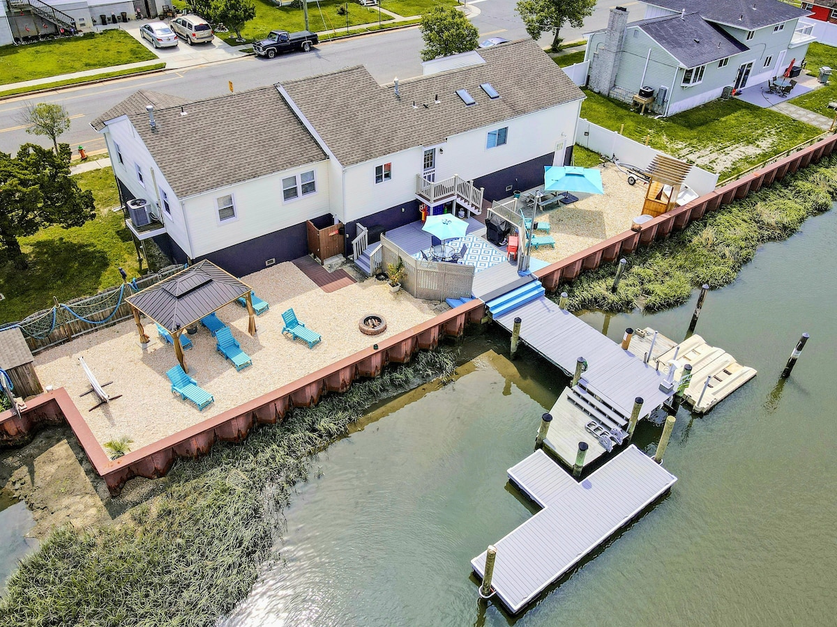 An aerial view captures the bayside getaway home with a spacious outdoor area featuring a gazebo, lounge chairs, and a pool. A private dock extends into the canal, providing access to the water. The surrounding yard is well-maintained, offering a relaxing atmosphere.