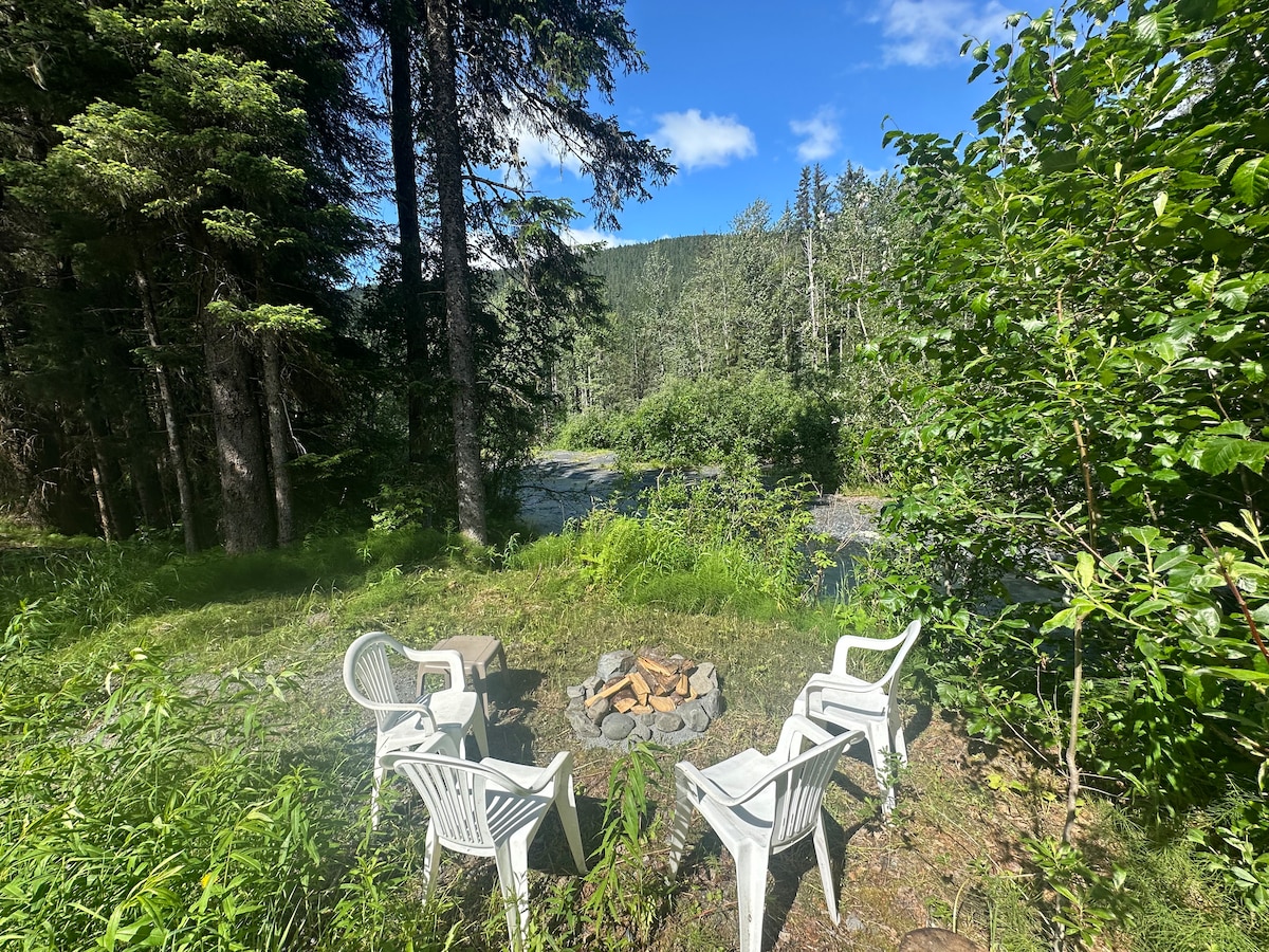 A serene outdoor seating area is surrounded by lush greenery, with four white chairs arranged around a stone fire pit. A gentle creek flows nearby, framed by tall trees and a clear blue sky overhead.