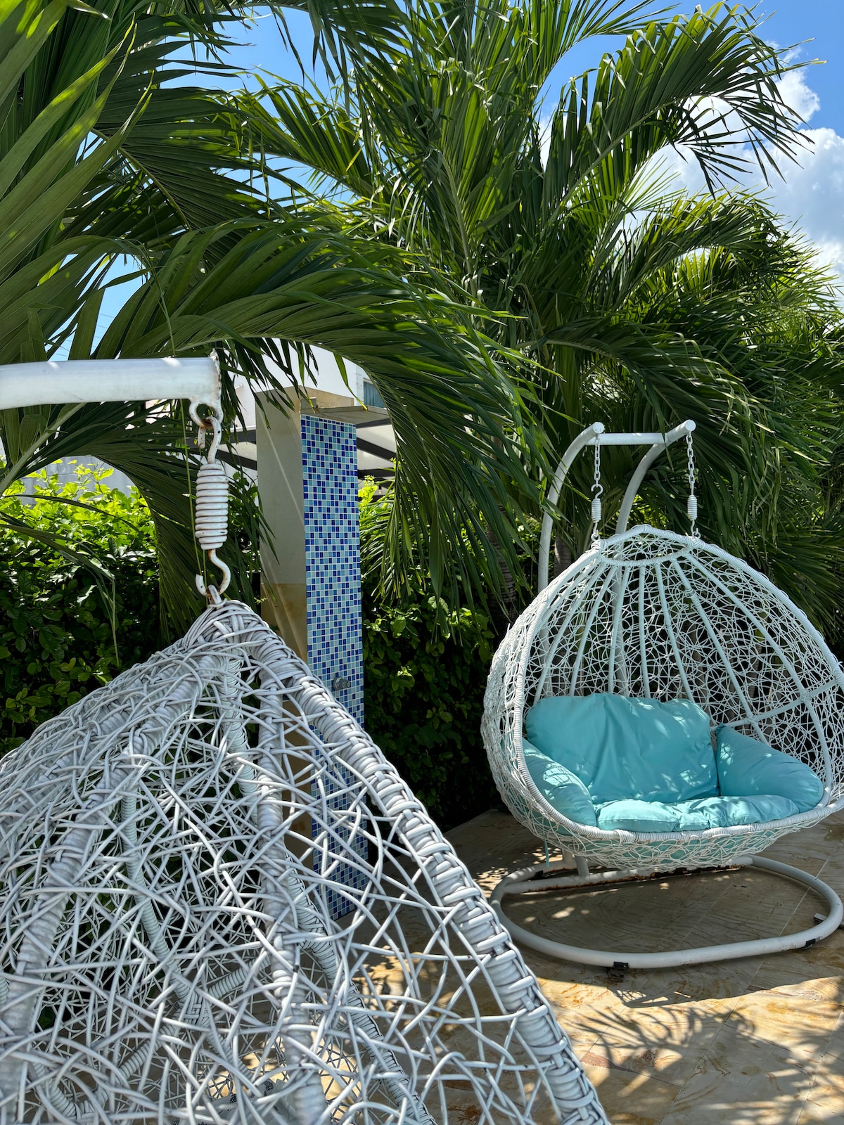 A pair of hanging chairs is positioned on a sunlit patio, surrounded by lush green palm foliage. One chair features a soft blue cushion, inviting relaxation. The area is complemented by a textured wall with a mosaic pattern in the background.