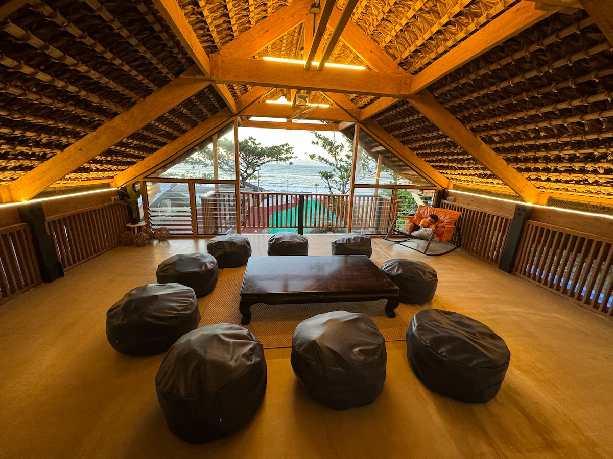 The loft area features a high wooden ceiling with decorative tiles and warm lighting. Multiple bean bag seating arrangements surround a low wooden table, while large windows offer views of the beach and natural surroundings.