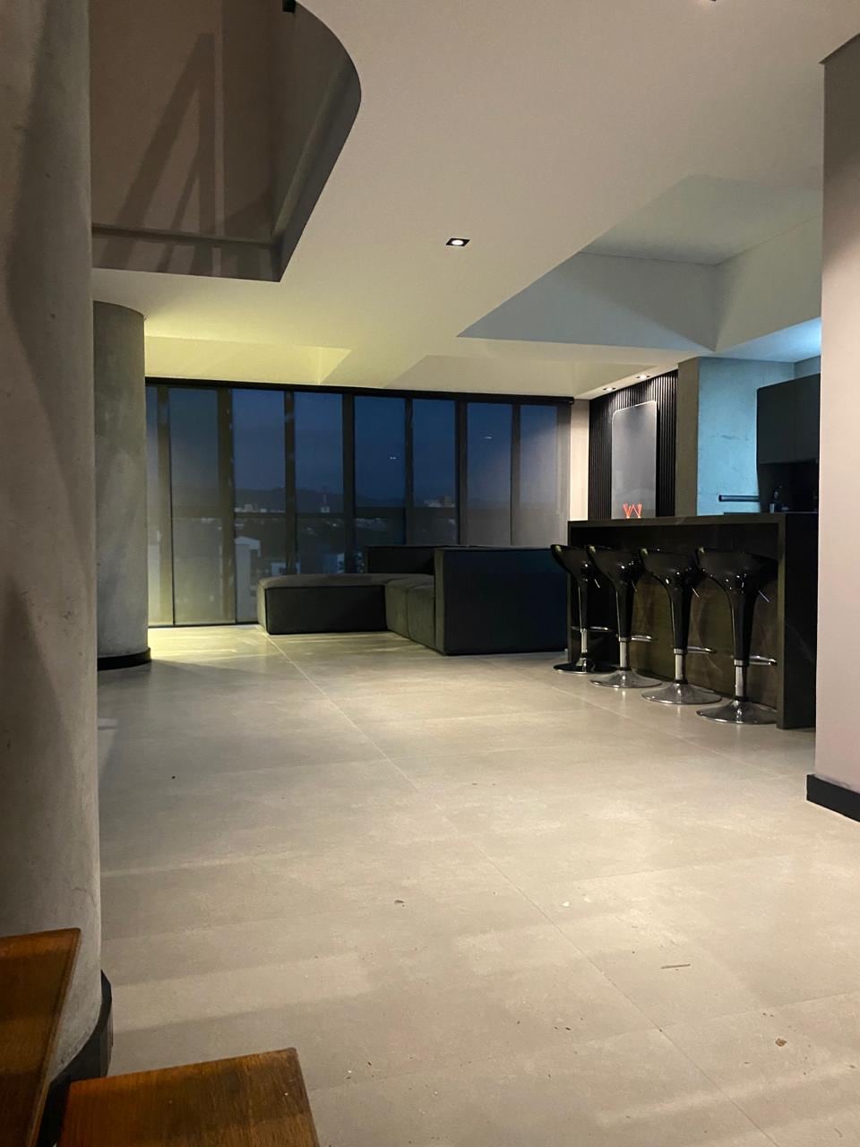 The open living area features modern architecture with sleek furnishings. Large floor-to-ceiling windows allow natural light to fill the space while revealing views of the city. A bar area with high stools complements the minimalist design, creating an inviting atmosphere.